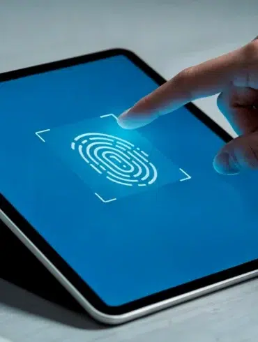 A hand touches a tablet screen displaying a large fingerprint icon on a blue background, symbolizing biometric security. The setting is minimal and modern.