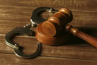 Metal handcuffs rest on a wooden surface next to a wooden gavel.