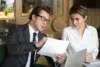 Man in a suit and glasses discusses a document with a woman in a white blouse. They are seated at a table with a laptop, focused and engaged.