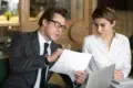 Man in a suit and glasses discusses a document with a woman in a white blouse. They are seated at a table with a laptop, focused and engaged.