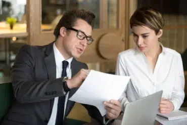 Man in a suit and glasses discusses a document with a woman in a white blouse. They are seated at a table with a laptop, focused and engaged.