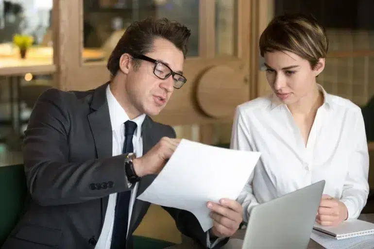 Man in a suit and glasses discusses a document with a woman in a white blouse. They are seated at a table with a laptop, focused and engaged.