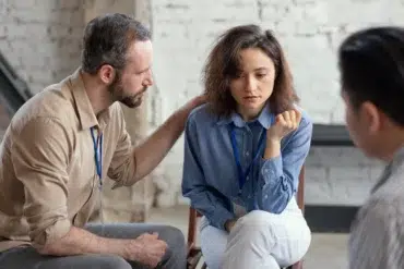 A man places a comforting hand on a distressed woman's shoulder during a group therapy session.
