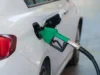 White car refueling at a gas station with a green fuel nozzle inserted into the fuel tank opening.