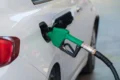 White car refueling at a gas station with a green fuel nozzle inserted into the fuel tank opening.