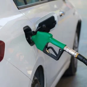 White car refueling at a gas station with a green fuel nozzle inserted into the fuel tank opening.