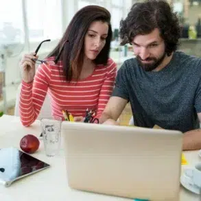 Two people intently work at a laptop in a bright office. A woman holds glasses and a man uses the laptop. Nearby are snacks, a tablet, and plants.