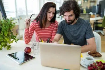 Two people intently work at a laptop in a bright office. A woman holds glasses and a man uses the laptop. Nearby are snacks, a tablet, and plants.