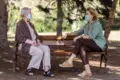 Two women wearing masks sit on a park bench, engaged in conversation. Sunlight filters through trees, creating a calm, peaceful atmosphere.