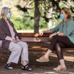 Two women wearing masks sit on a park bench, engaged in conversation. Sunlight filters through trees, creating a calm, peaceful atmosphere.