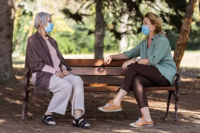 Two women wearing masks sit on a park bench, engaged in conversation. Sunlight filters through trees, creating a calm, peaceful atmosphere.