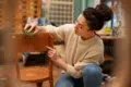 Woman in a cozy sweater refurbishes a wooden chair with a sanding sponge in a workshop. The setting is warm and creative, suggesting a DIY project.