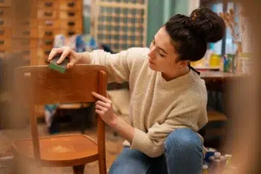 Woman in a cozy sweater refurbishes a wooden chair with a sanding sponge in a workshop. The setting is warm and creative, suggesting a DIY project.