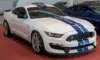 White Ford Mustang Shelby GT350 with blue racing stripes displayed indoors on a red floor alongside other cars.