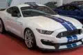 White Ford Mustang Shelby GT350 with blue racing stripes displayed indoors on a red floor alongside other cars.