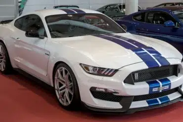 White Ford Mustang Shelby GT350 with blue racing stripes displayed indoors on a red floor alongside other cars.