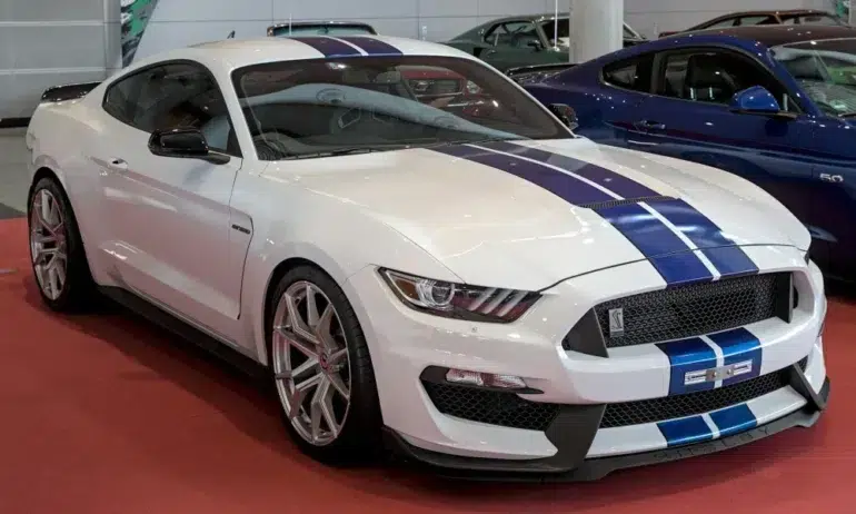 White Ford Mustang Shelby GT350 with blue racing stripes displayed indoors on a red floor alongside other cars.