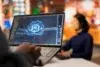 A person's hands are on a laptop displaying a glowing blue brain with "AI" in the center, as another person in the background wears a headset.