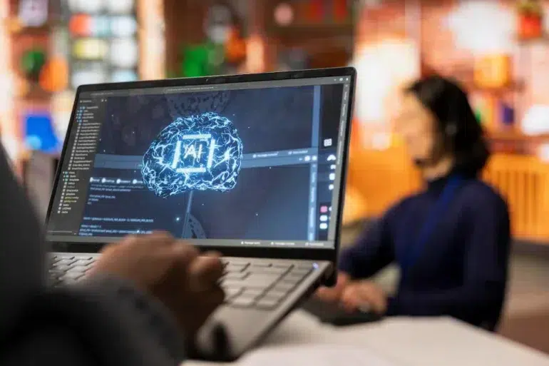 A person's hands are on a laptop displaying a glowing blue brain with "AI" in the center, as another person in the background wears a headset.