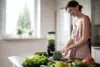 Woman preparing fresh broccoli in a bright kitchen with various fruits and vegetables and a blender with green smoothie on the counter.