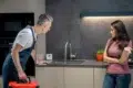 A plumber in overalls, carrying a red toolbox, discusses with a woman in a kitchen. She looks concerned, gesturing towards the sink. Modern setting, tense mood.