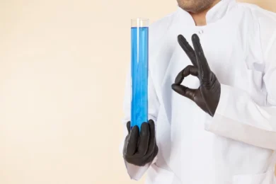A scientist in a white coat and black gloves holds a large test tube filled with blue liquid, making an "OK" gesture. The tone is professional and precise.