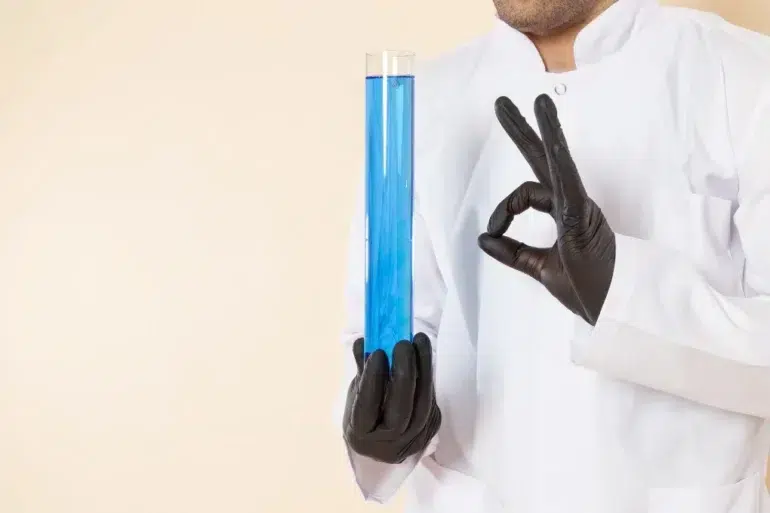 A scientist in a white coat and black gloves holds a large test tube filled with blue liquid, making an "OK" gesture. The tone is professional and precise.