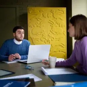 A man and woman engaged in discussion at a table, with a laptop and papers. Background features a yellow panel with animal motifs, creating a focused atmosphere.