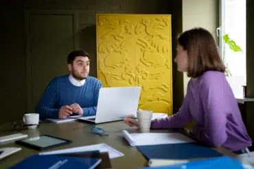 A man and woman engaged in discussion at a table, with a laptop and papers. Background features a yellow panel with animal motifs, creating a focused atmosphere.