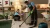 Two people in blue shirts and overalls clean a modern kitchen. One vacuums the floor with green gloves, while the other wipes a cabinet, surrounded by plants.