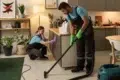 Two people in blue shirts and overalls clean a modern kitchen. One vacuums the floor with green gloves, while the other wipes a cabinet, surrounded by plants.