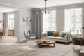 A bright, modern living room with a cream sofa, blue and beige pillows, and a wooden coffee table. Large windows, grey curtains, and green plants add a fresh touch.