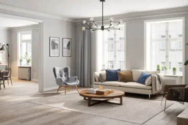 A bright, modern living room with a cream sofa, blue and beige pillows, and a wooden coffee table. Large windows, grey curtains, and green plants add a fresh touch.