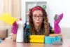 Woman in glasses and a red headband holding cleaning supplies, including a yellow cloth and steel wool.
