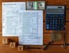 Tax documents, calculator, magnifying glass, and $100 bills on a wooden table. Wooden blocks spell "TAX," suggesting financial calculation.