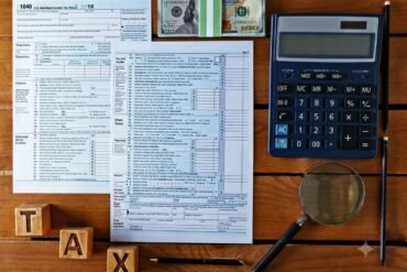 Tax documents, calculator, magnifying glass, and $100 bills on a wooden table. Wooden blocks spell "TAX," suggesting financial calculation.