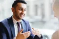 A man in a suit, smiling and gesturing while talking to another person in an office setting. The atmosphere is professional and engaging.