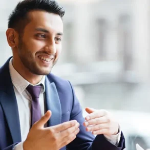 A man in a suit, smiling and gesturing while talking to another person in an office setting. The atmosphere is professional and engaging.