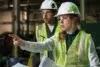 Two construction workers in hard hats and vests. The woman points decisively while holding papers, conveying focus and teamwork in a construction setting.