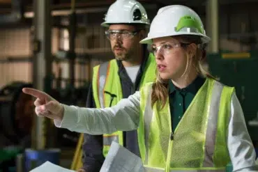 Two construction workers in hard hats and vests. The woman points decisively while holding papers, conveying focus and teamwork in a construction setting.