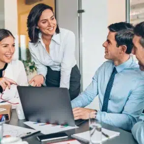 A group of four colleagues in formal attire are gathered around a laptop in an office, smiling and engaged in a lively discussion, conveying teamwork.
