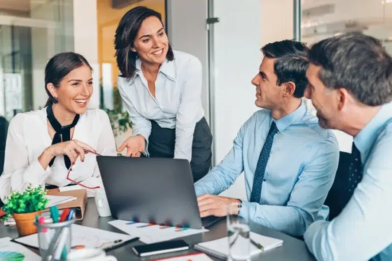 A group of four colleagues in formal attire are gathered around a laptop in an office, smiling and engaged in a lively discussion, conveying teamwork.