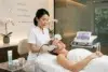 Aesthetician in a white uniform uses a device for facial treatment on a relaxed woman lying on a spa bed. The room is calm, with soft lighting and orchids.