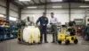 Two men in a workshop examine industrial equipment, including a large white tank and a yellow machine. One holds a tablet, discussing details.