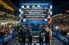 Four racing drivers stand on a podium with a crowd cheering in the background and a large screen displaying "The Importance of Reliability in Crypto Payment Infrastructure" at the 24 Hours of Le Mans.