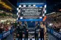 Four racing drivers stand on a podium with a crowd cheering in the background and a large screen displaying "The Importance of Reliability in Crypto Payment Infrastructure" at the 24 Hours of Le Mans.
