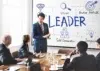 A person in a suit presents to a group in a conference room. The whiteboard displays the word "LEADER" surrounded by diagrams with traits like "Vision" and "Inspiration."