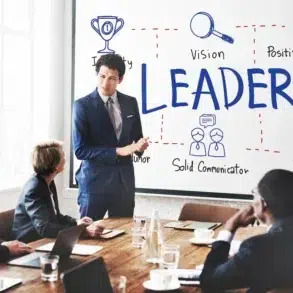 A person in a suit presents to a group in a conference room. The whiteboard displays the word "LEADER" surrounded by diagrams with traits like "Vision" and "Inspiration."