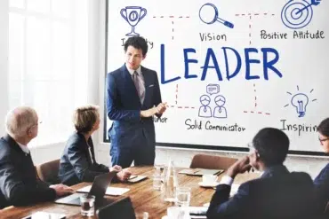A person in a suit presents to a group in a conference room. The whiteboard displays the word "LEADER" surrounded by diagrams with traits like "Vision" and "Inspiration."