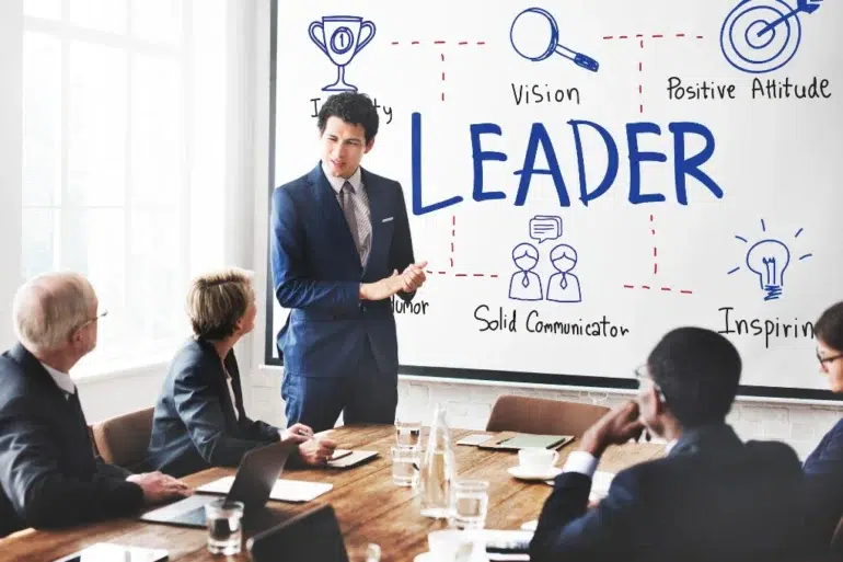 A person in a suit presents to a group in a conference room. The whiteboard displays the word "LEADER" surrounded by diagrams with traits like "Vision" and "Inspiration."